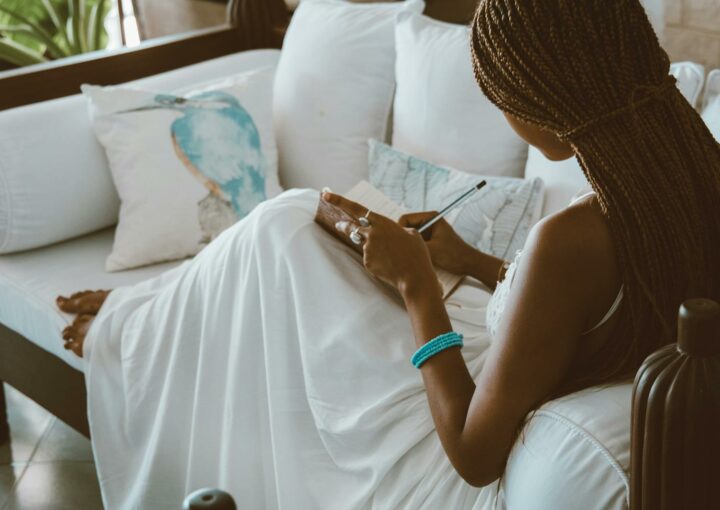 An African American woman in a white dress writing in a notebook while sitting on a sofa indoors.