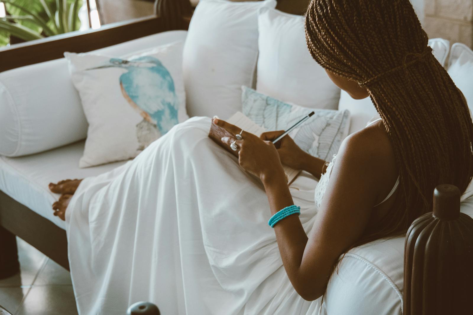 An African American woman in a white dress writing in a notebook while sitting on a sofa indoors.