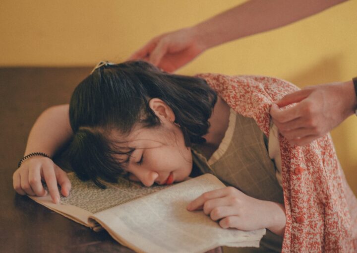 A young woman falls asleep on a book as someone covers her with a blanket.