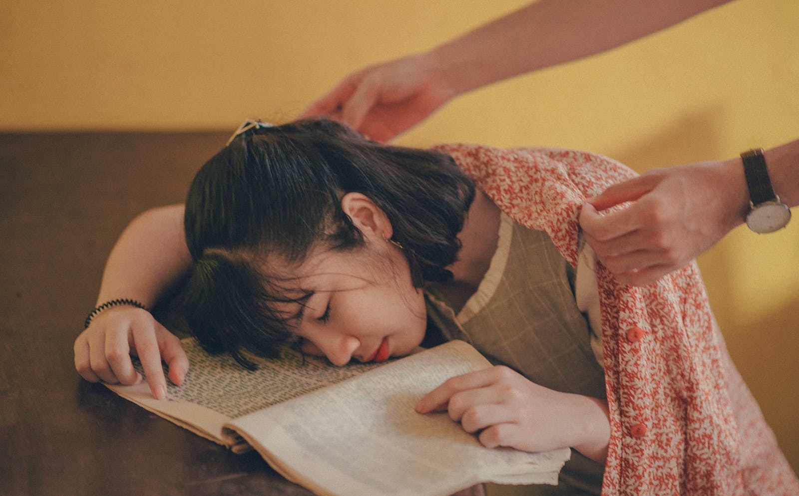 A young woman falls asleep on a book as someone covers her with a blanket.