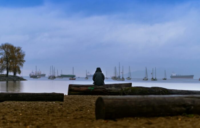 A lone figure sits on a sandy beach in Vancouver, gazing at boats and the calm ocean under a dusky sky.