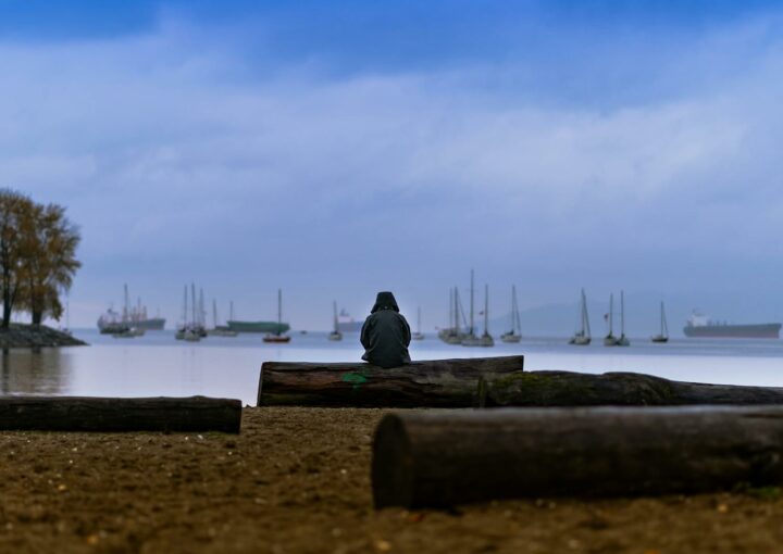 A lone figure sits on a sandy beach in Vancouver, gazing at boats and the calm ocean under a dusky sky.