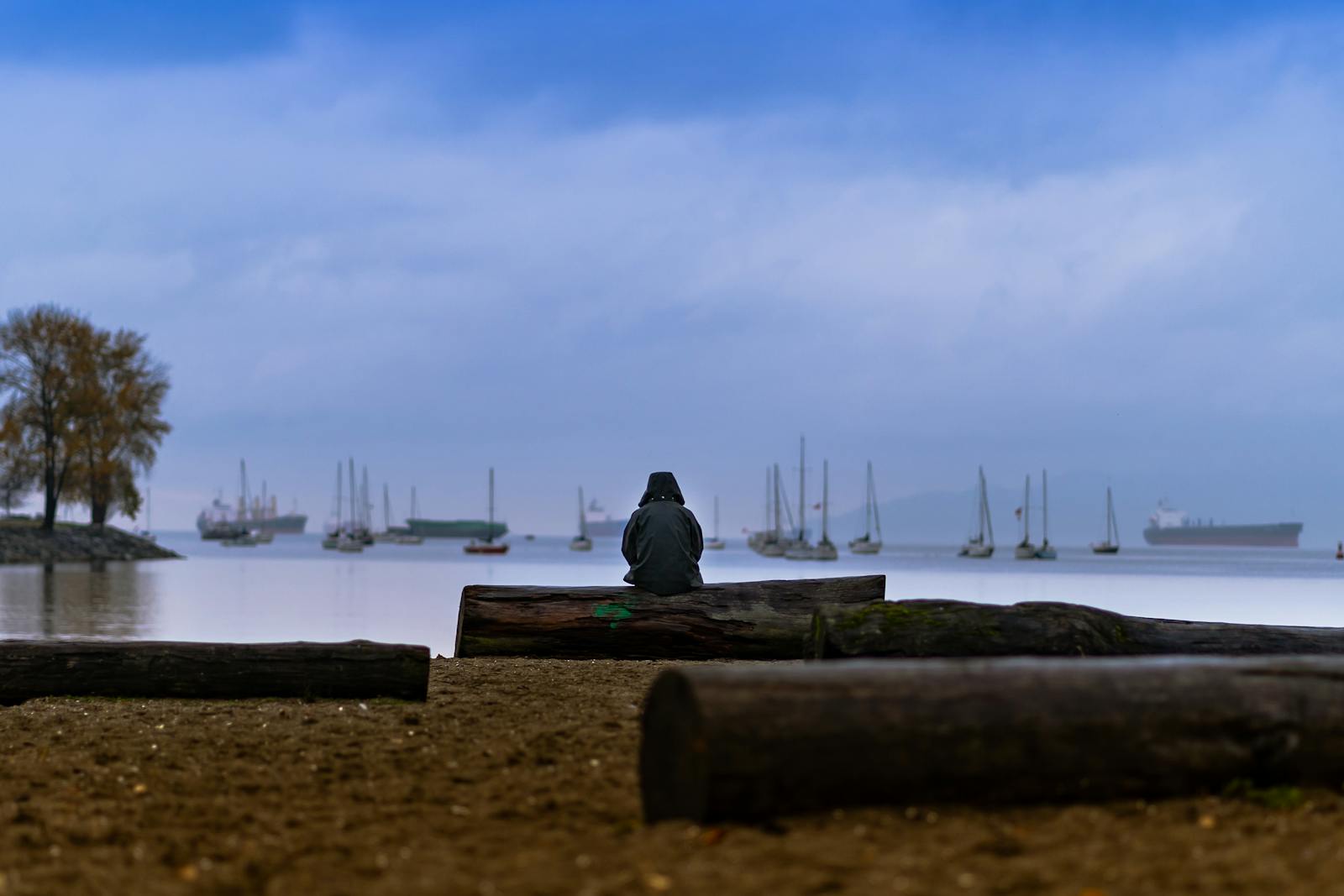 A lone figure sits on a sandy beach in Vancouver, gazing at boats and the calm ocean under a dusky sky.