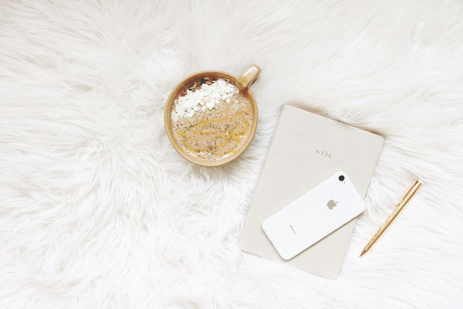 Minimalist flat lay of coffee, smartphone, notebook, and pen on white fur.