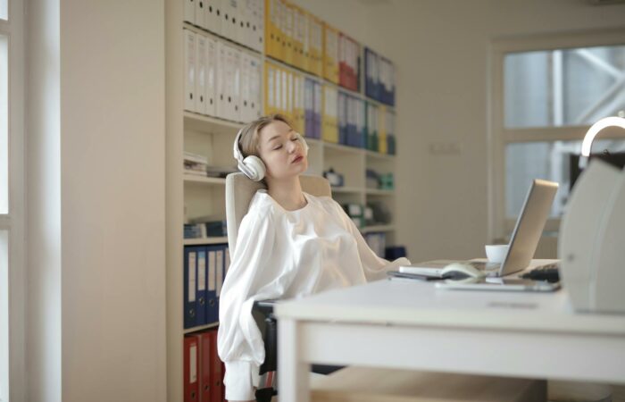 Woman in white shirt relaxing at office desk with headphones, appearing restful.