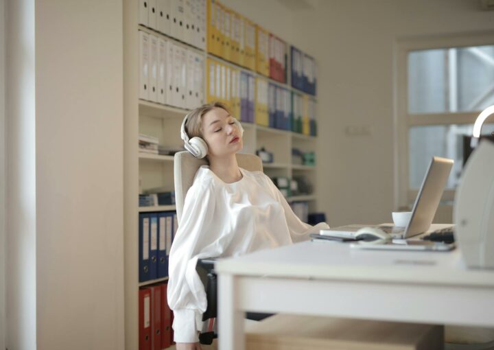 Woman in white shirt relaxing at office desk with headphones, appearing restful.