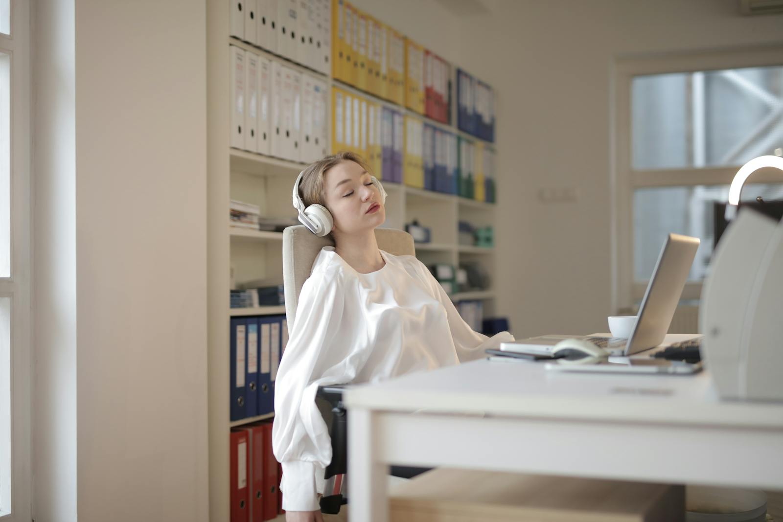 Woman in white shirt relaxing at office desk with headphones, appearing restful.