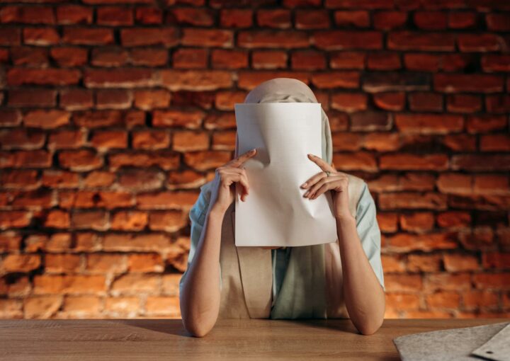 Individual at desk holds paper, obscuring face against a textured brick wall.