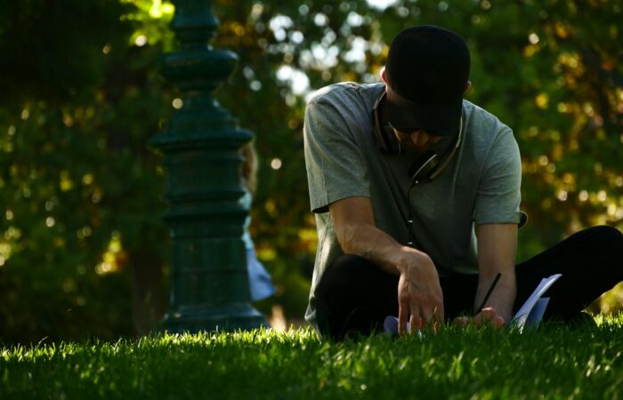 selective focus photography of man sitting on grass and writing on notebook during daytime