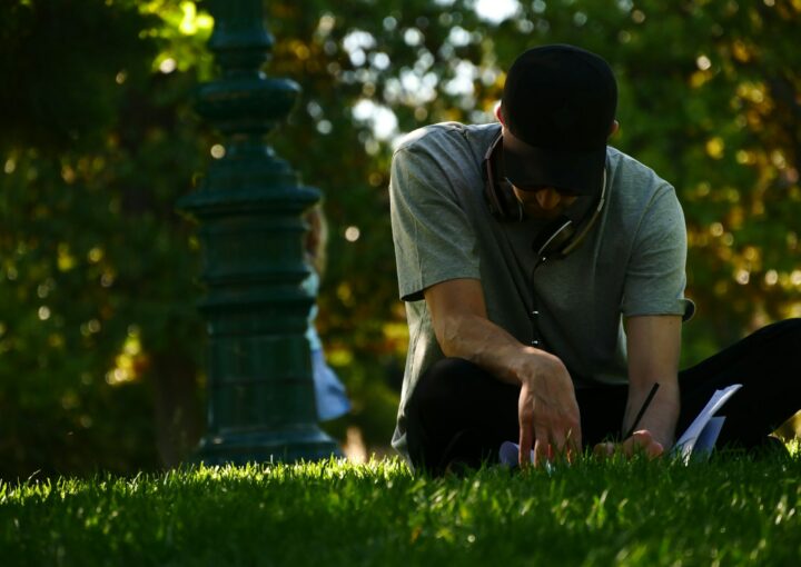 selective focus photography of man sitting on grass and writing on notebook during daytime