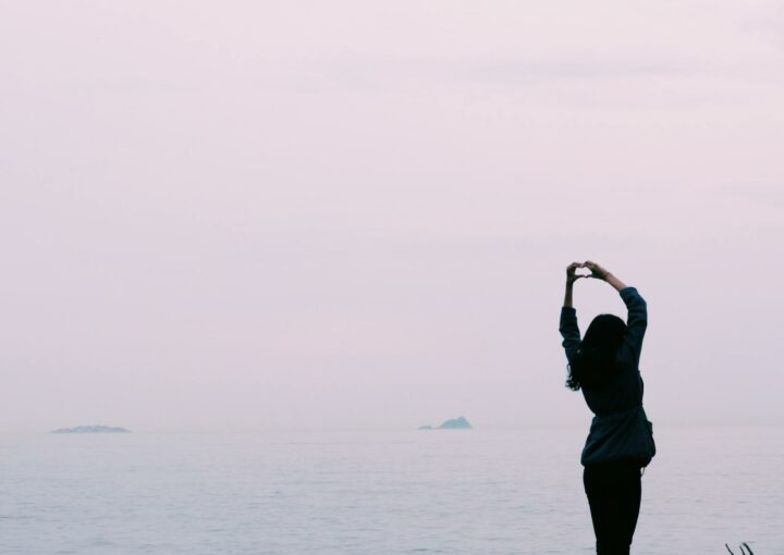 A silhouette of a woman making a heart shape with her hands by the calm sea during sunset.