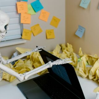 A skeletal figure works at a desk with a laptop and scattered papers, symbolizing burnout.